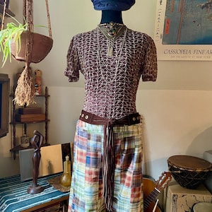 May include: A mannequin displays a vintage outfit. It includes a dark blue cap, a brown lace top, a plaid skirt with a brown belt, and layered necklaces. The outfit is styled with a bohemian aesthetic.