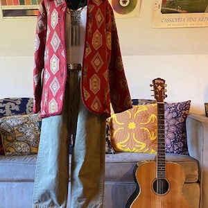 May include: A red patterned jacket, a white top, and olive-green pants are displayed on a mannequin. A wooden acoustic guitar leans against a sofa with decorative pillows. The background includes art prints.