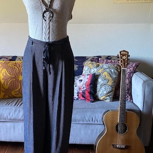 May include: A mannequin displays a light gray knit top with a cable knit design and a black necklace. Below, a pair of navy blue pinstripe pants with a drawstring waist. A guitar leans against a couch with decorative pillows.