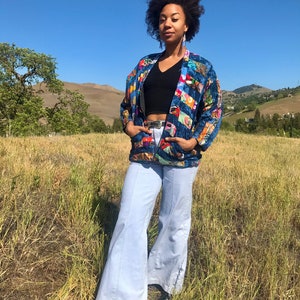 Pode incluir: Uma mulher vestindo uma jaqueta de patchwork azul e multicolorida com um top preto e calças boca de sino azul claro. Ela está em pé em um campo de grama alta.