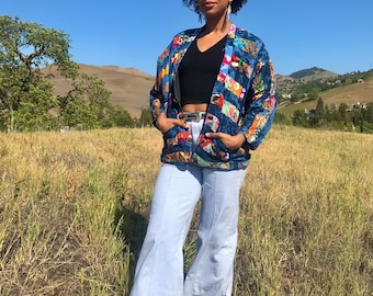 May include: A woman wearing a blue and multi-colored patchwork jacket with a black top and light blue bell-bottom pants. She is standing in a field of tall grass.