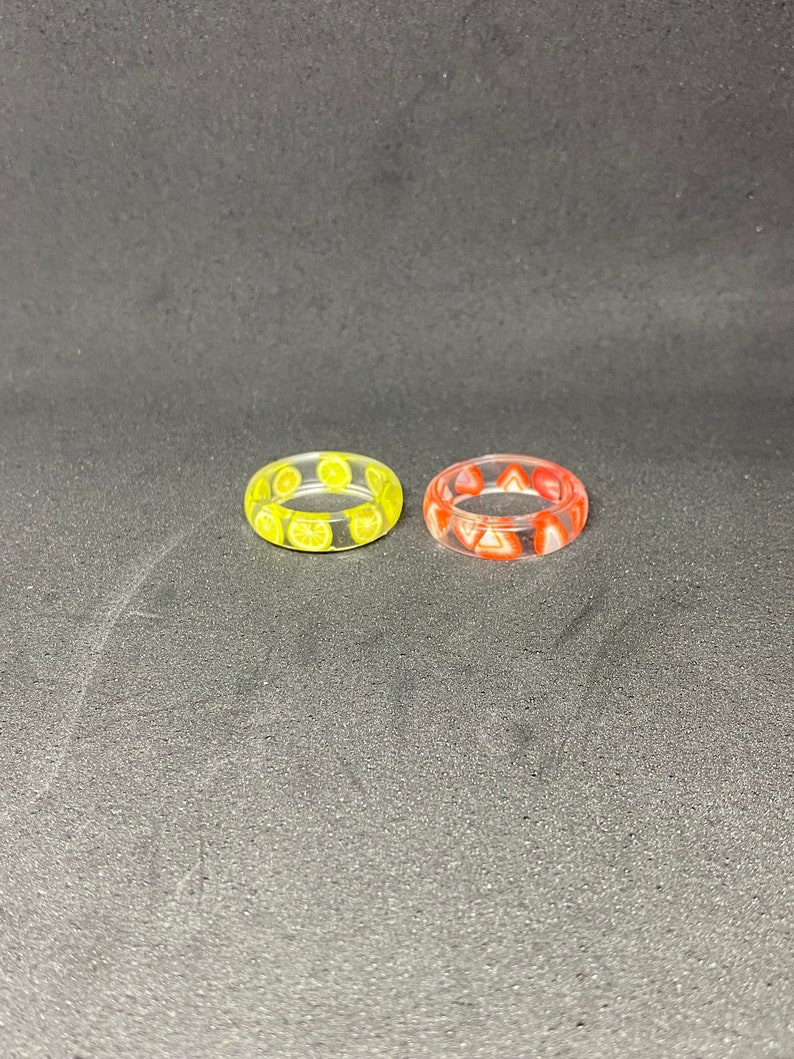 May include: Two clear resin rings with fruit designs. One ring has a yellow lemon design and the other has a red strawberry design.