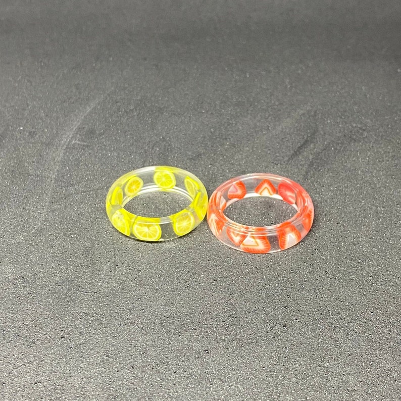 May include: Two clear resin rings with embedded fruit slices. One ring has yellow lemon slices and the other has red strawberry slices.