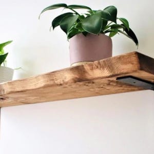 May include: A rustic, wooden shelf with a natural finish, supported by black metal brackets. A potted plant with large, green leaves sits on the shelf, adding a touch of nature. The shelf is mounted against a white wall, creating a contrast.