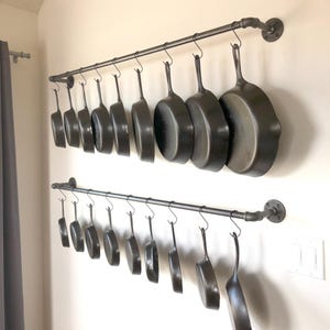 May include: Two rows of cast iron pans hang from a metal pipe rack on a wall. The pans are various sizes and shapes, with long handles and a dark gray color. The rack is made of black metal pipes and fittings.