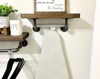 May include: A wall-mounted wooden shelf with black metal pipe brackets holding an ironing board with a beige and white striped cover. A sign above reads: dirty... will you do me? yours truly, the laundry. A white HOME mug and green plant sit nearby.