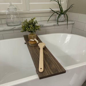 May include: A dark brown wooden bathtub tray with a light brown wooden brush and a round brush with a white strap. The tray holds a small green plant and a small box. The bathtub is white.