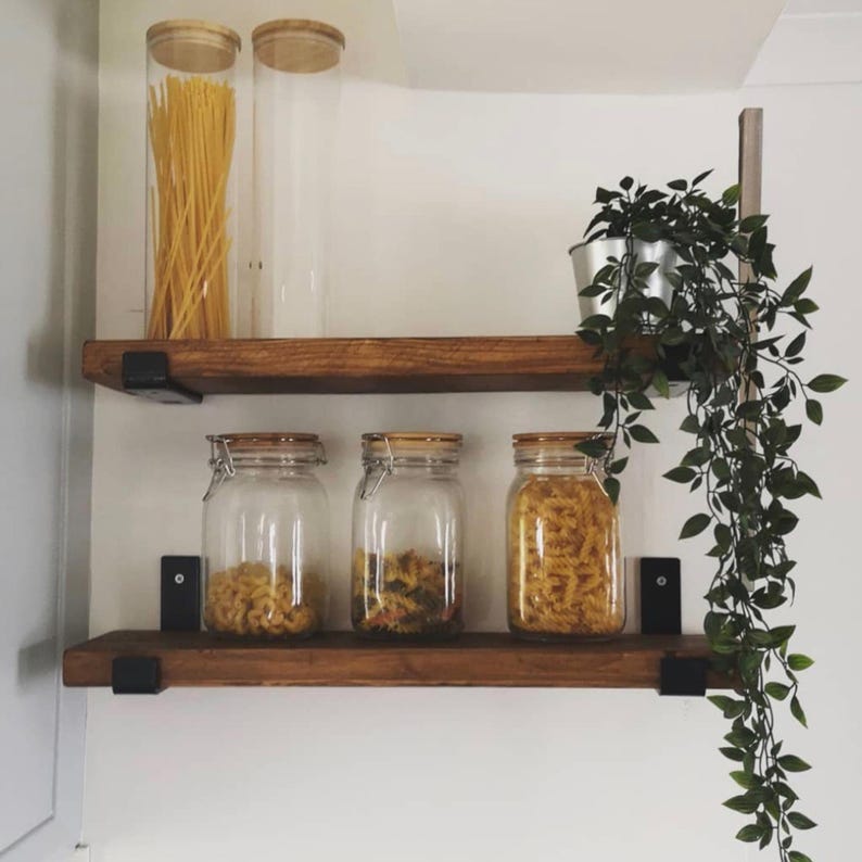 May include: Wooden shelves with black metal brackets hold glass jars filled with pasta and other dry goods. Two tall jars with bamboo lids stand on the top shelf. A small potted plant hangs from the right side.