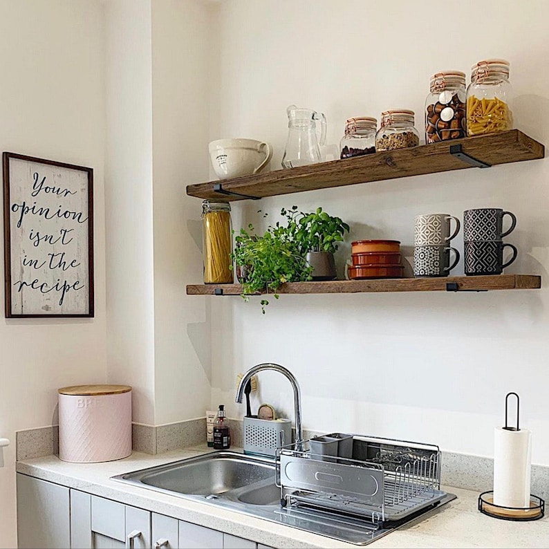 May include: A kitchen scene with wooden shelves holding jars of food, mugs, and plants. A framed sign reads "Your opinion isn't in the recipe." A pink bread bin and a stainless steel sink are also visible.