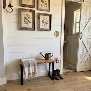 May include: A wooden bench with black metal legs features a cream and gray plaid throw blanket draped over one side. A small metal watering can sits on the bench surface. The bench is positioned against a white shiplap wall with four framed botanical prints.