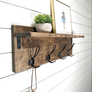 May include: A wooden wall shelf with four black metal hooks and a small potted plant. The shelf has a dark brown stain and is mounted on a white wall. A framed print with a gold frame is on the shelf.