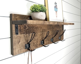 Könnte beinhalten: Ein wandmontiertes Holzregal mit dunkelbraun gebeizter Oberfläche und vier schwarzen Metallhaken. Auf dem oberen Regalbrett steht eine kleine grüne Topfpflanze neben einem gerahmten Bild einer Person, die einen Stapel Wäsche trägt.