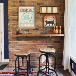 May include: A rustic home bar setup with a brick wall backdrop. Features include a wooden shelf with bottles and glasses, two adjustable bar stools, and framed artwork with the text "Whiskey Map" and "Superstition Black Label Whiskey."