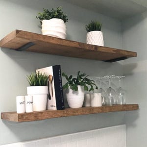 May include: Two rustic wooden shelves mounted on a light blue wall. The top shelf holds white ceramic pots with greenery. The bottom shelf displays a book, small white pots, and wine glasses. The shelves are supported by black metal brackets.