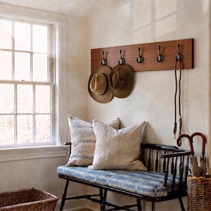 May include: A wooden wall-mounted coat rack with five dark metal hooks holding two felt hats and a brown rope leash. Below, a black wooden bench features a blue and white plaid cushion and two textured throw pillows, with umbrellas nearby.