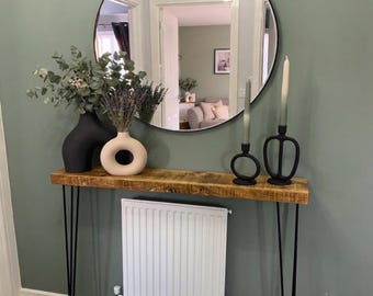 May include: A console table with a thick, rectangular wooden top and four thin black metal hairpin legs. The table is positioned against a sage green wall, featuring a large round mirror, a black vase, a beige donut vase, and two black candle holders.