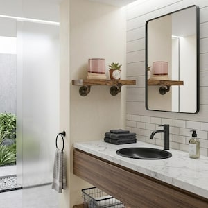 May include: A bathroom vanity featuring a white marble countertop, a black oval sink, and a black faucet. Above, a rectangular mirror with a black frame hangs next to a wooden wall shelf supported by dark metal pipe brackets holding a plant and candle.