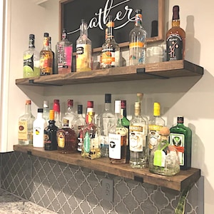 May include: Two wooden shelves displaying various liquor bottles. The shelves are supported by black metal brackets and mounted on a white wall. A chalkboard sign with the word "gather" in cursive is above the top shelf. The shelves are above a tiled countertop.