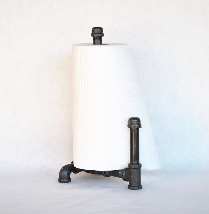May include: Black metal pipe paper towel holder with a white paper towel roll. The holder has a three-legged base and a pipe extending up to hold the paper towel roll.