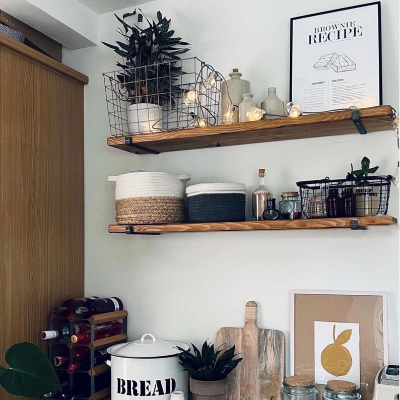 May include: A kitchen scene with wooden shelves displaying various decorative items. A wire basket holds a plant, and a framed "Brownie Recipe" poster hangs above. Other items include jars, bottles, and a bread bin. A wine rack and cutting boards are also visible.