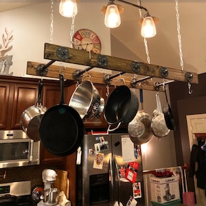 May include: A rustic, wooden pot rack suspended from the ceiling by chains. The rack features metal pipes and hooks, holding various pots and pans. Above, three pendant lights with glass shades illuminate the kitchen. A decorative clock is visible on the wall.