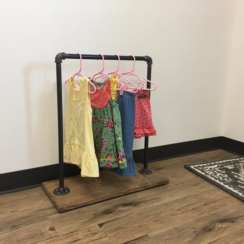 May include: A clothing rack constructed from black industrial pipes mounted on a rectangular wooden base. Four pink plastic hangers are suspended from the top bar, holding a yellow dress, a floral print dress, blue pants, and a pink patterned top.