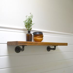 May include: A rectangular wooden shelf with a natural brown finish, supported by two dark gray metal pipe brackets. On the shelf sits a small amber glass bottle containing a green plant and a stack of four small bowls in varying wood and dark tones.