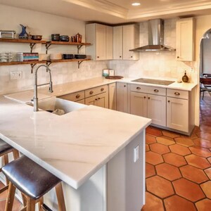 Pu&ograve; includere: Una cucina moderna con mobili bianchi, un piano di lavoro in marmo e una cappa in acciaio inossidabile. Mensole aperte espongono stoviglie e decorazioni. Il pavimento &egrave; piastrellato con piastrelle esagonali color terracotta. Un rubinetto in acciaio inossidabile &egrave; sopra il lavandino.
