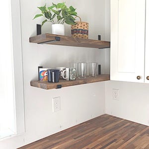 May include: Two wooden shelves with black metal brackets are mounted on a white wall. The top shelf holds a potted plant and a woven basket. The bottom shelf displays mugs and clear drinking glasses. The shelves are in a kitchen setting.