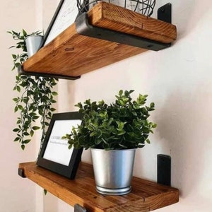 May include: Two rustic wooden shelves with black metal brackets are mounted on a white wall. The shelves hold decorative items, including a framed print with text, a metal bucket with a plant, and hanging greenery. The shelves are made of stained wood.