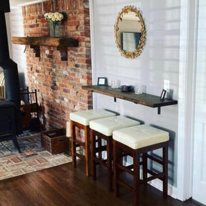 May include: Three wooden bar stools with cream-colored padded seats are positioned in front of a wall-mounted wooden shelf. A gold-framed oval mirror hangs above the shelf. The background features a brick wall and a wooden floor.