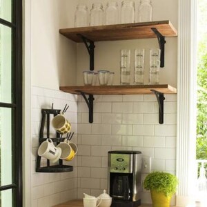 Pu&ograve; includere: Una scena di cucina con due mensole in legno con staffe in metallo nero, che contengono barattoli di vetro e bicchieri. Sotto, una macchina per il caff&egrave;, un portabicchieri con tazze e una piccola pianta in un vaso giallo sono visibili. Lo sfondo comprende piastrelle bianche e una finestra.