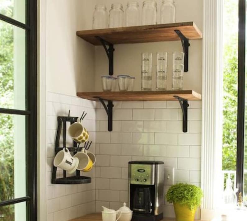 Pu&ograve; includere: Una scena di cucina con due mensole in legno con staffe in metallo nero, che contengono barattoli di vetro e bicchieri. Sotto, una macchina per il caff&egrave;, un portabicchieri con tazze e una piccola pianta in un vaso giallo sono visibili. Lo sfondo comprende piastrelle bianche e una finestra.
