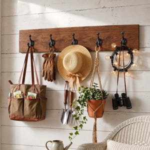May include: A rectangular wooden wall rack with five black metal hooks. Items hanging include a brown canvas tote bag, a tan leather glove, a straw hat with a ribbon, two metal gardening tools, a potted plant in a macrame hanger, and string lights.