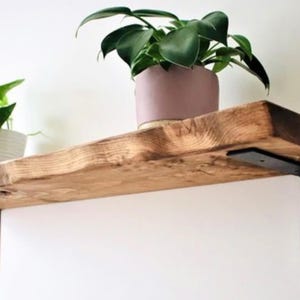 May include: A rustic wooden shelf with a natural wood grain finish. A potted plant with green leaves sits on the shelf. The shelf is supported by black metal brackets and mounted on a white wall. Home decor.