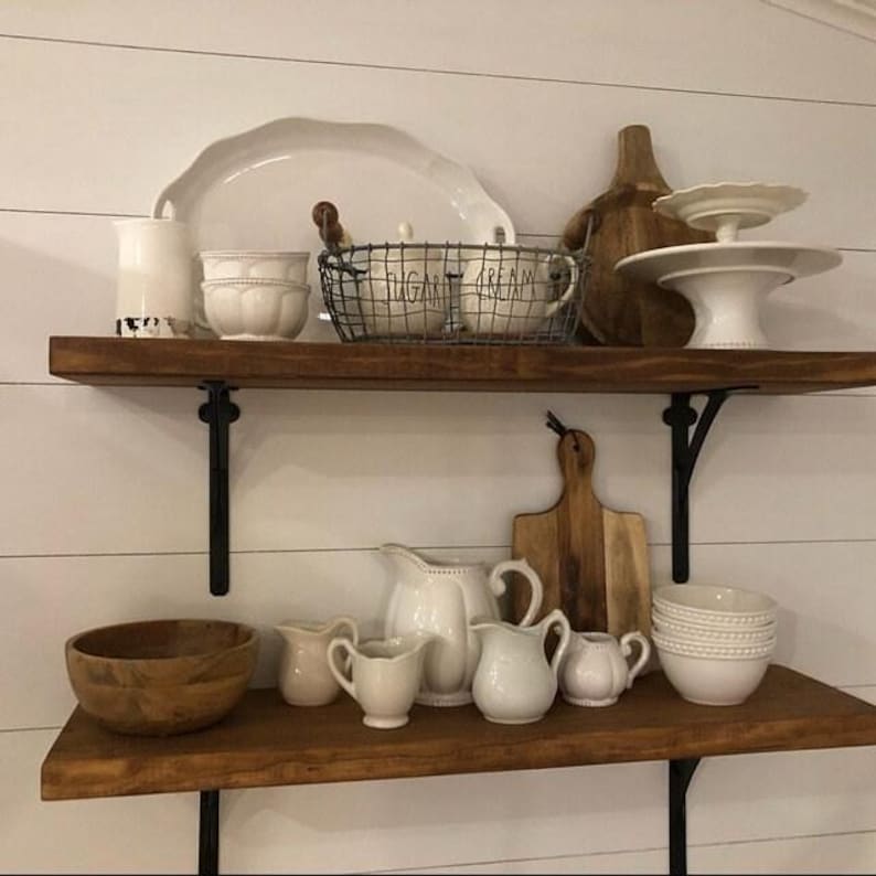 Pu&ograve; includere: Esposizione di stoviglie da cucina su mensole rustiche in legno. Gli oggetti includono ciotole, brocche e piatti di servizio in ceramica bianca. Un cestino di filo metallico contiene tazze con le scritte "ZUCCHERO" e "PANNA".