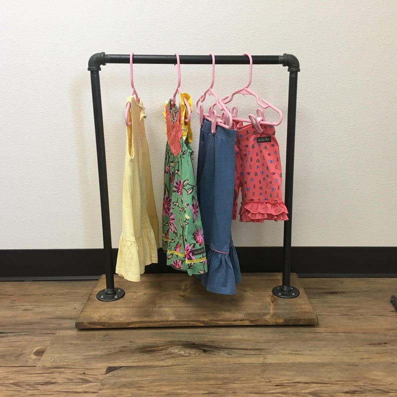 May include: A black industrial pipe clothing rack with a rectangular wooden base. Four pink plastic hangers are suspended from the top bar, holding a yellow dress, a green floral dress, blue pants, and pink patterned shorts with ruffled hems.
