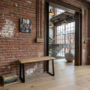 May include: A rectangular wooden bench with a natural light brown finish and black metal U-shaped legs. The bench is positioned against a red brick wall, featuring a smooth top surface and a sturdy frame design that provides a stable seating base.