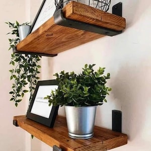 May include: Two rustic wooden shelves with black metal brackets are mounted on a wall. The shelves hold a framed print, a metal bucket with a green plant, and hanging greenery. The shelves are made of stained wood.