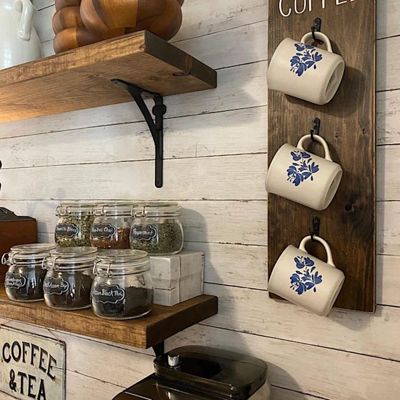 Pu&ograve; includere: Una scena di cucina rustica con mensole in legno, barattoli di spezie e un portabicchieri da caff&egrave;. Tre tazze bianche con disegni floreali blu sono appese a una tavola di legno scuro. Sono visibili barattoli di spezie e t&egrave;.