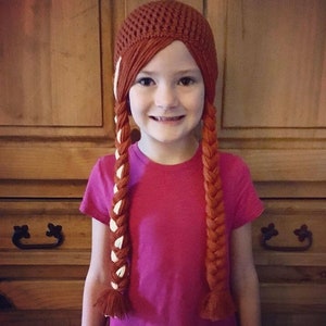 May include: A crocheted brown hat with attached long, braided orange hair, accented with white strands. The hat is worn by a person wearing a pink t-shirt. The background is a wooden cabinet.