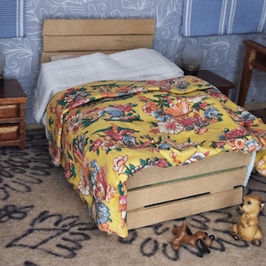 May include: Miniature bedroom scene featuring a wooden bed with a yellow floral patterned comforter. The bed is made of wooden crates. A small wooden nightstand with a lamp sits beside the bed. A laptop is on a small table. Two small animal figurines are on the floor.