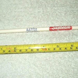 Op de afbeelding: Een witte potlood met het woord "BATTER" in blauw en "Sunbeam" in rood gedrukt. Een gele rolmaat ligt naast het potlood. De potlood is ongeveer 19 cm lang.