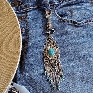Peut inclure: Un accessoire porte-clés en métal argenté présentant un motif filigrane orné avec une pierre ovale de couleur turquoise au centre. L'objet comprend un motif fleur de lys, un fermoir mousqueton et plusieurs chaînes argentées pendantes terminées par de petites perles turquoise.