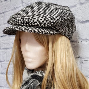 Peut inclure: Casquette de style gavroche à motif pied-de-poule noir et blanc. La casquette a une couronne arrondie et une courte visière rigide. Elle est présentée sur une tête de mannequin avec des cheveux blonds et une écharpe noire et blanche.