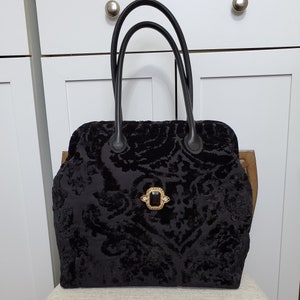 Close up image of black jacquard velvet carpet bag created with upholstery fabric in a damask pattern. The bag features top handles in black leather. bag is decorated with a vintage gold tone brooch framing a sparkling black faux gemstone.