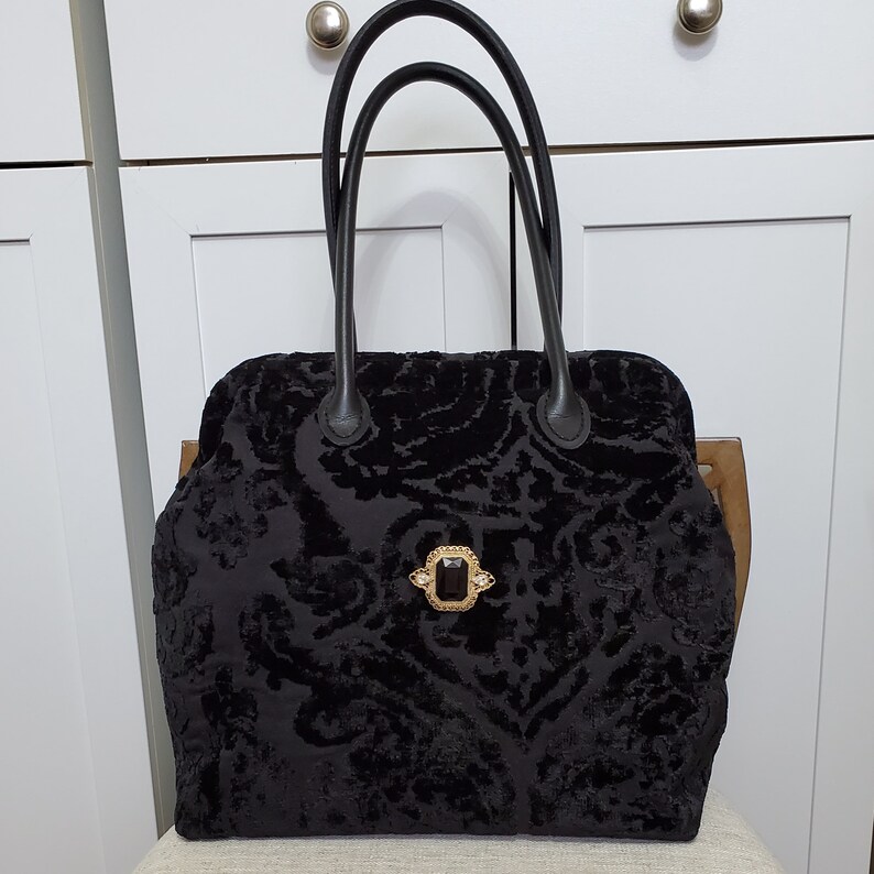 Close up image of black jacquard velvet carpet bag created with upholstery fabric in a damask pattern. The bag features top handles in black leather. bag is decorated with a vintage gold tone brooch framing a sparkling black faux gemstone.