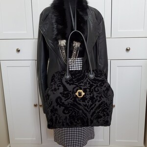Image of black jacquard velvet carpet bag displayed on a mannequin. Bag is created with upholstery fabric. The bag features top handles in black leather. Bag is decorated with a vintage gold tone brooch framing a sparkling black faux gemstone.
