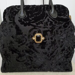 Close up image of black jacquard velvet carpet bag created with upholstery fabric in a damask pattern. The bag features top handles in black leather. bag is decorated with a vintage gold tone brooch framing a sparkling black faux gemstone.