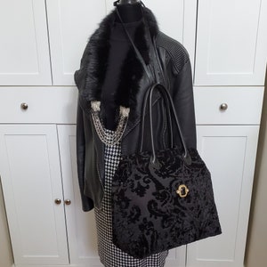 Image of black jacquard velvet carpet bag displayed on a mannequin. Bag is created with upholstery fabric. The bag features top handles in black leather. Bag is decorated with a vintage gold tone brooch framing a sparkling black faux gemstone.